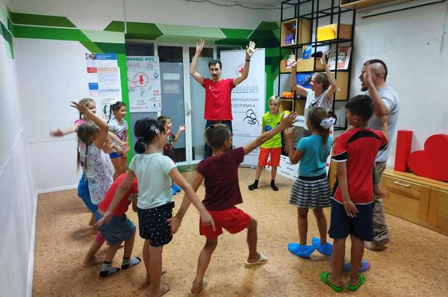 Children are standing in a circle in a big room and are learning tricks from the clowns (spinning plates).